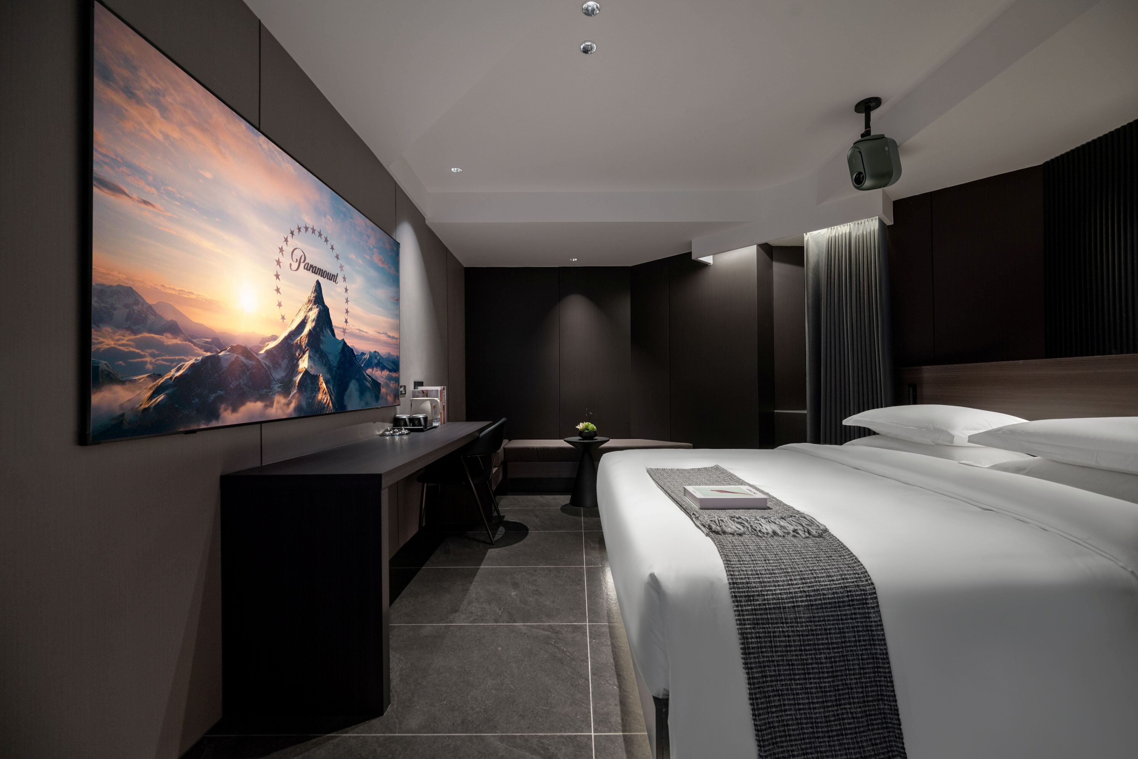Una camera d'albergo moderna con una grande TV a schermo piatto montata a parete, un letto rifatto con cura, una scrivania con sedia e un tavolino con una pianta.