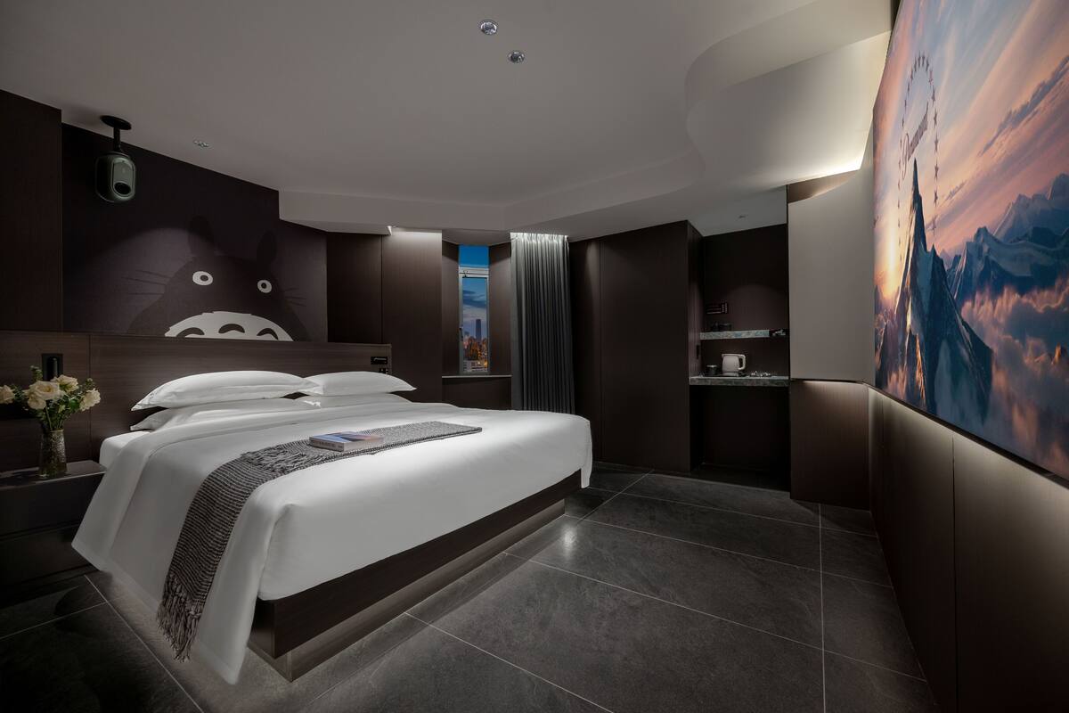 A modern hotel room with a large bed, a wall-mounted camera, a painting, and a view of the sky.