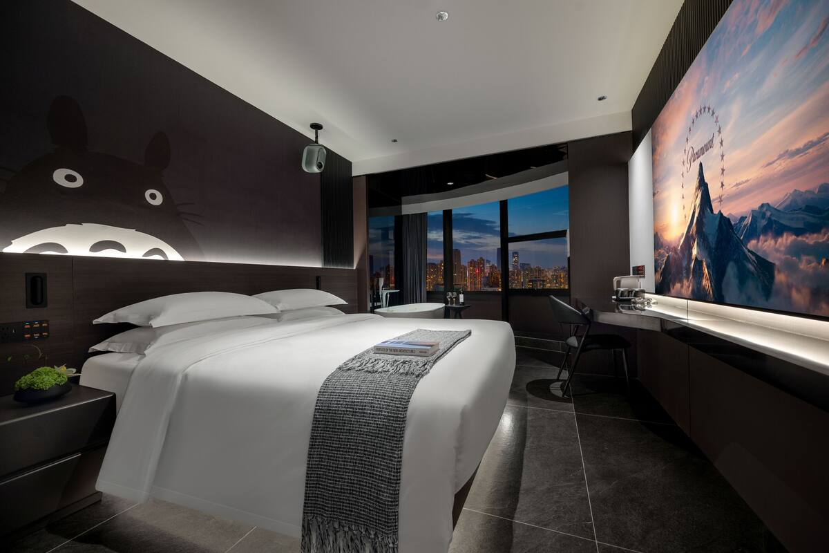 A modern hotel room with a large bed, a desk, a city view, and a mural on the wall.