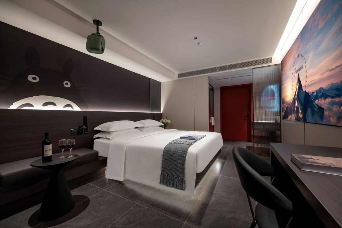 A modern hotel room with a large wall mural, a bed with a white comforter, a desk with a chair, and a small table with a bottle and glasses.
