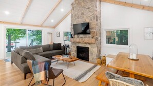 A spacious living room with a stone fireplace, a large sofa, a wooden dining table, and a ceiling fan. - Cedar Sauna, Cedar Hot Tub, Lakeside Fire Pit, Private Beach | La Dolce Muskoka (Bracebridge)