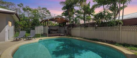 A swimming pool with a curved edge, surrounded by a fence, and a covered patio area with chairs.