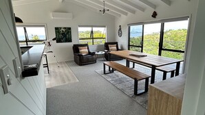 A spacious living area with a large window, a ceiling fan, and a dining table. - Te Puawaitanga Beach House - The Blossoming of Something New. (Tutukaka)