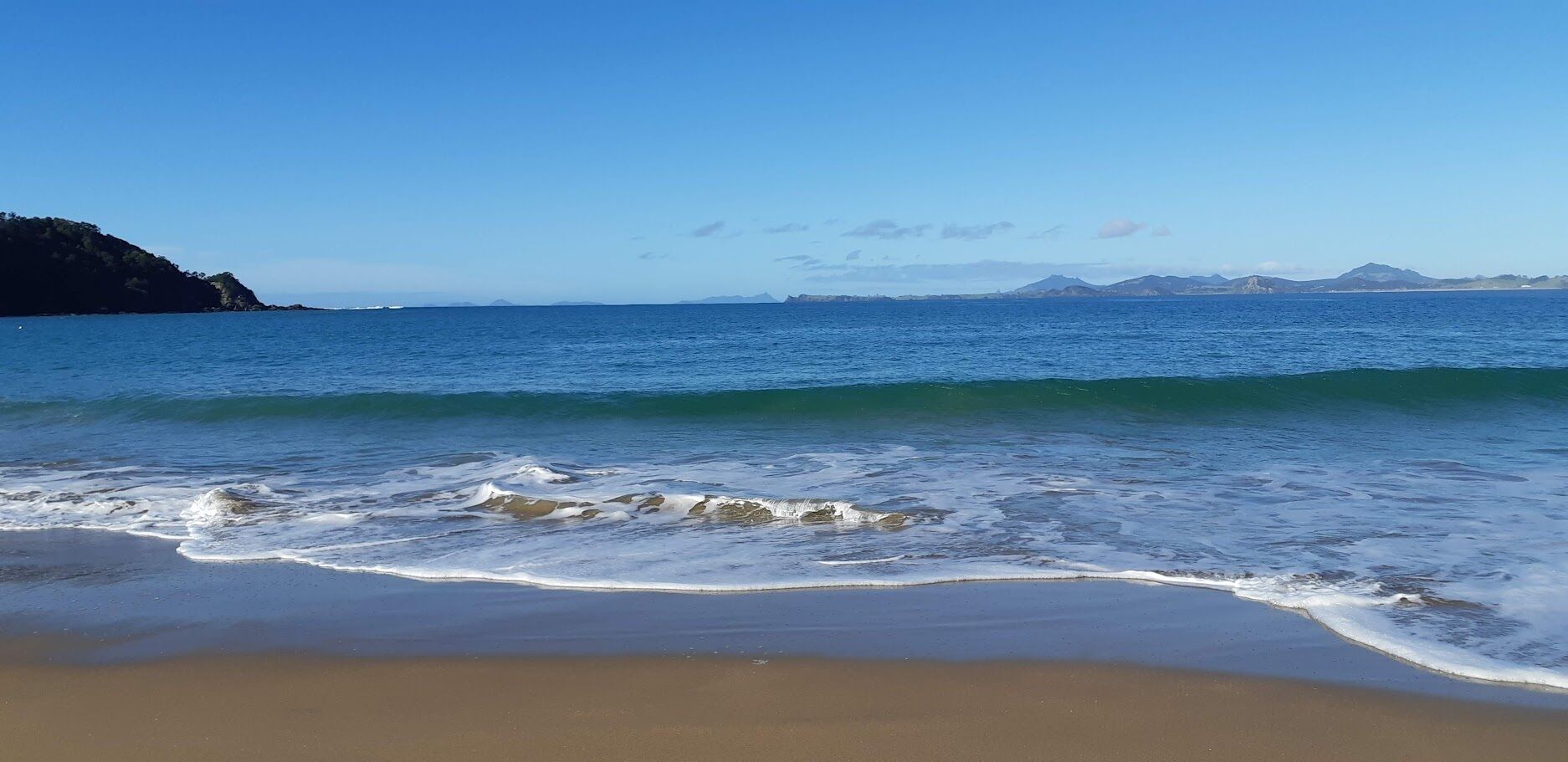 A beach with waves gently lapping onto the shore, a clear sky, and distant mountains.