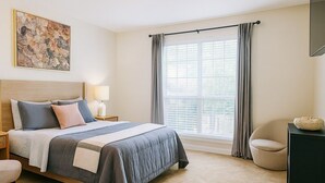 Une chambre à coucher comprenant un lit, une table de chevet, une chaise, un bureau, une fenêtre avec des rideaux et un tableau accroché au mur.