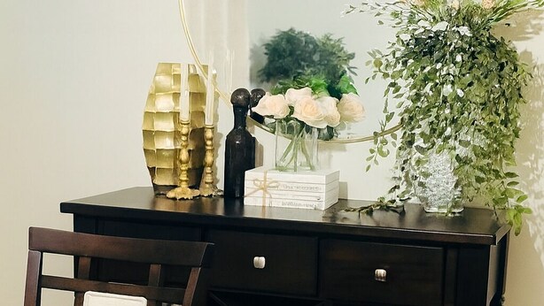 Un bureau en bois foncé, surmonté d’un miroir à cadre doré, un vase avec des fleurs blanches et une plante décorative.