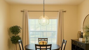 Un coin repas comprenant une table ronde, quatre chaises, un luminaire suspendu, une plante en pot et une fenêtre avec des stores.