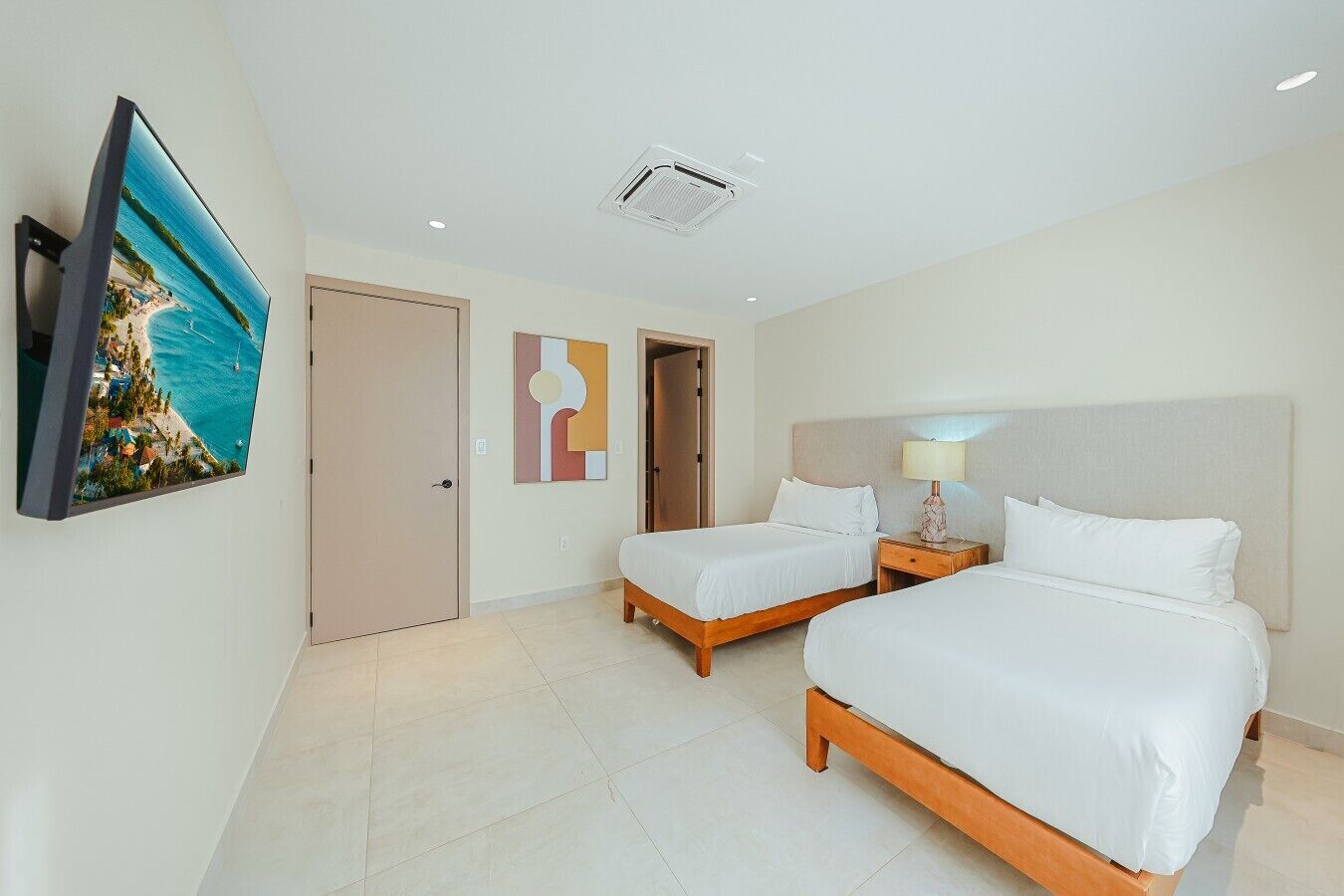 Quarto de hotel com duas camas, uma televisão, uma ventilação no teto e uma porta.