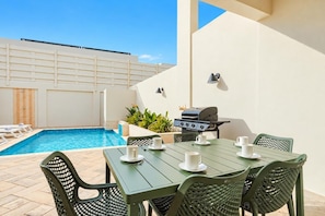 Zona de piscina con mesa para cuatro, una parrilla y sillas de descanso.
