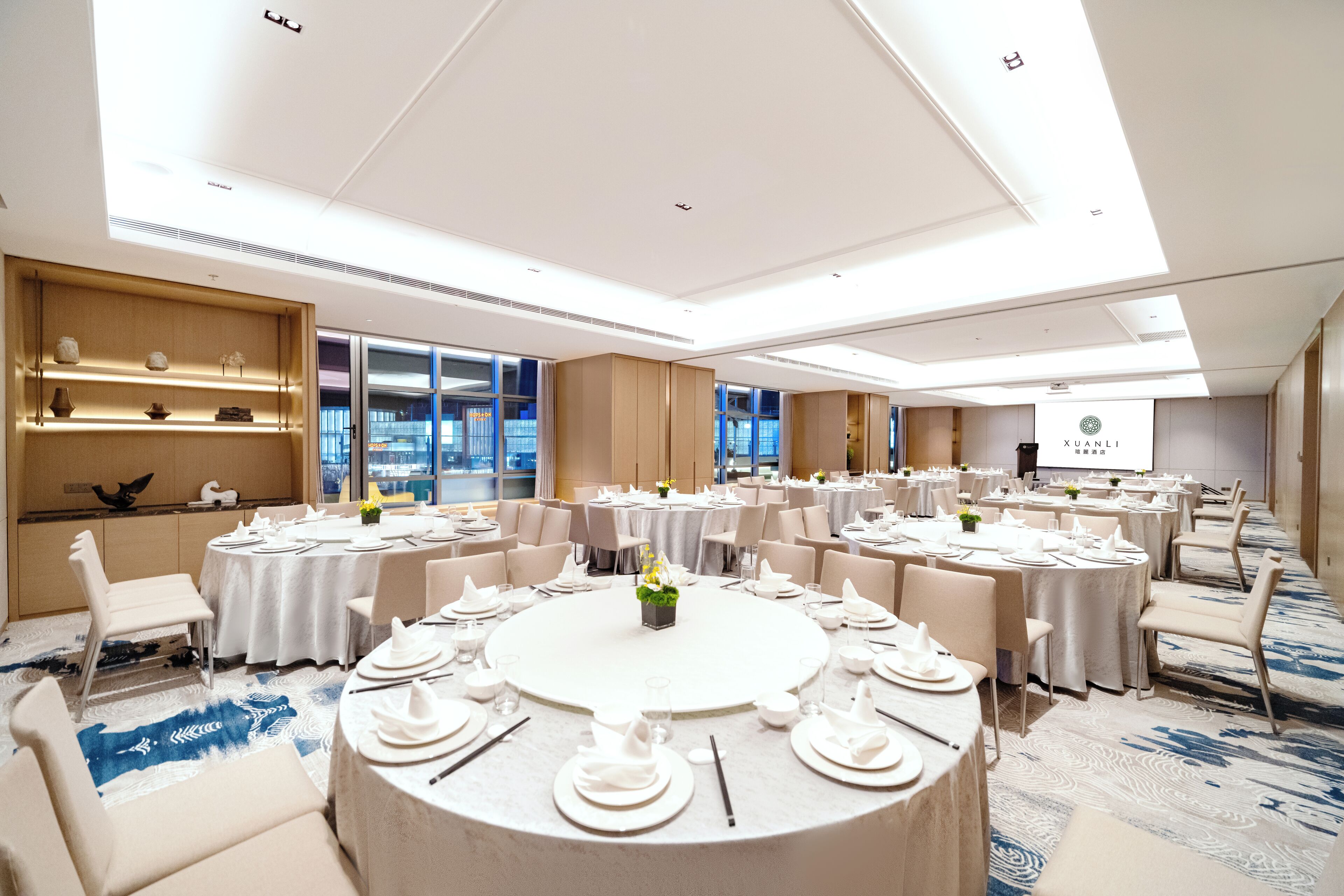 A spacious conference room with multiple round tables set for dining, equipped with white tablecloths, plates, and cutlery.