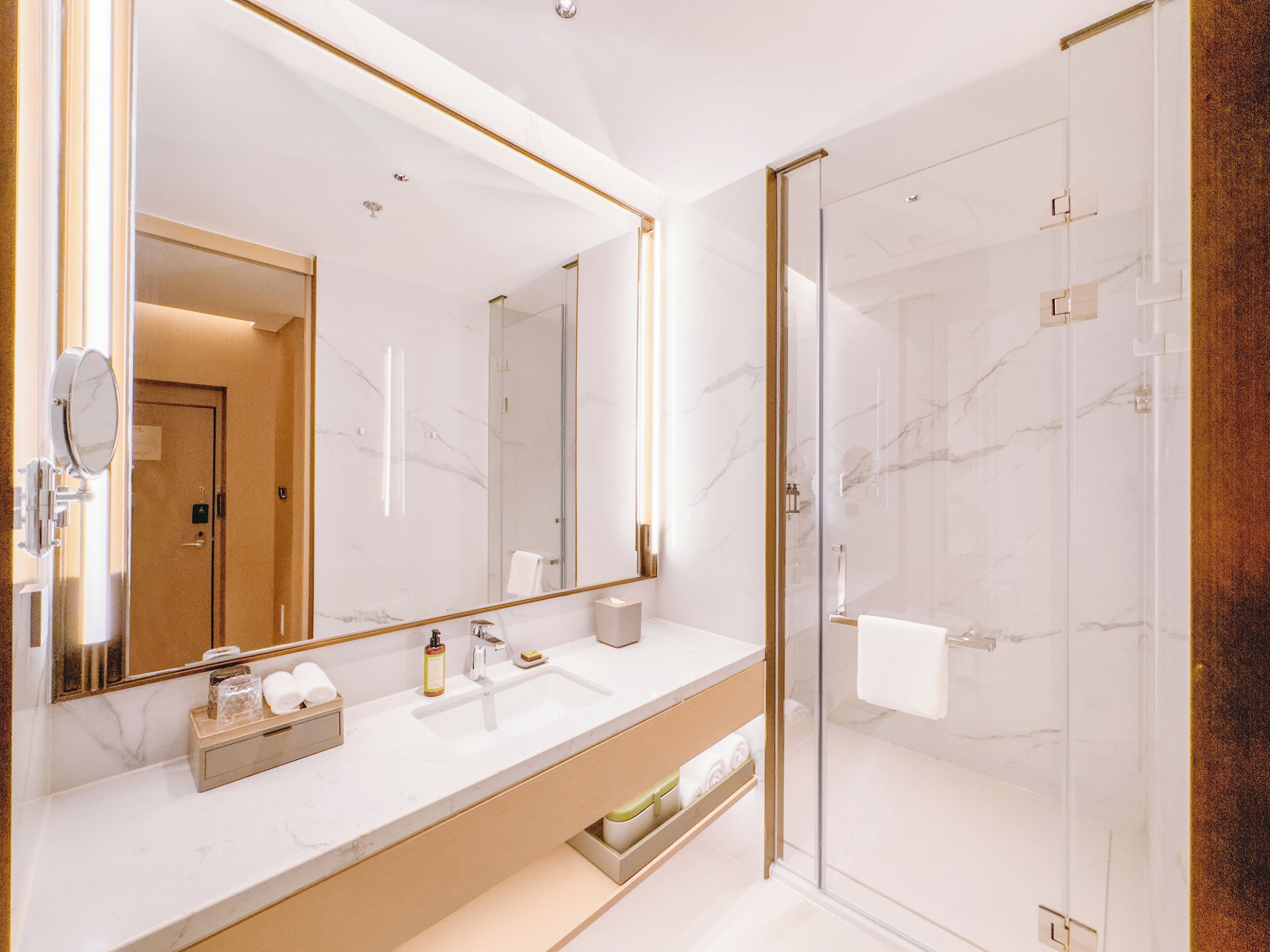 A modern bathroom with a large mirror, marble countertops, a glass shower enclosure, and a wooden vanity.