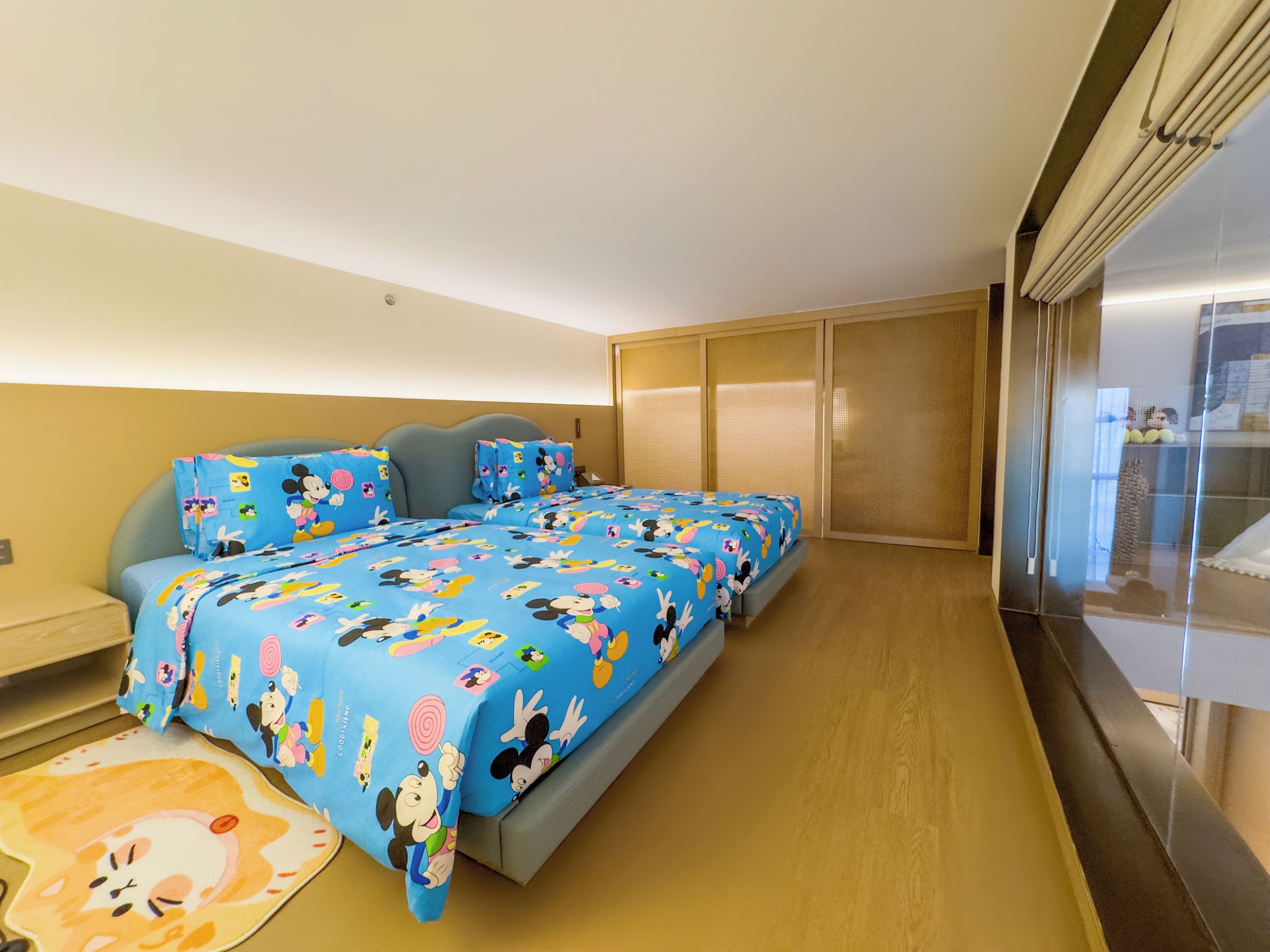 Une chambre d’hôtel avec un grand lit recouvert d’une couvre-lit aux motifs de Mickey Mouse, une petite table de chevet, une commode et une fenêtre donnant sur l’extérieur.