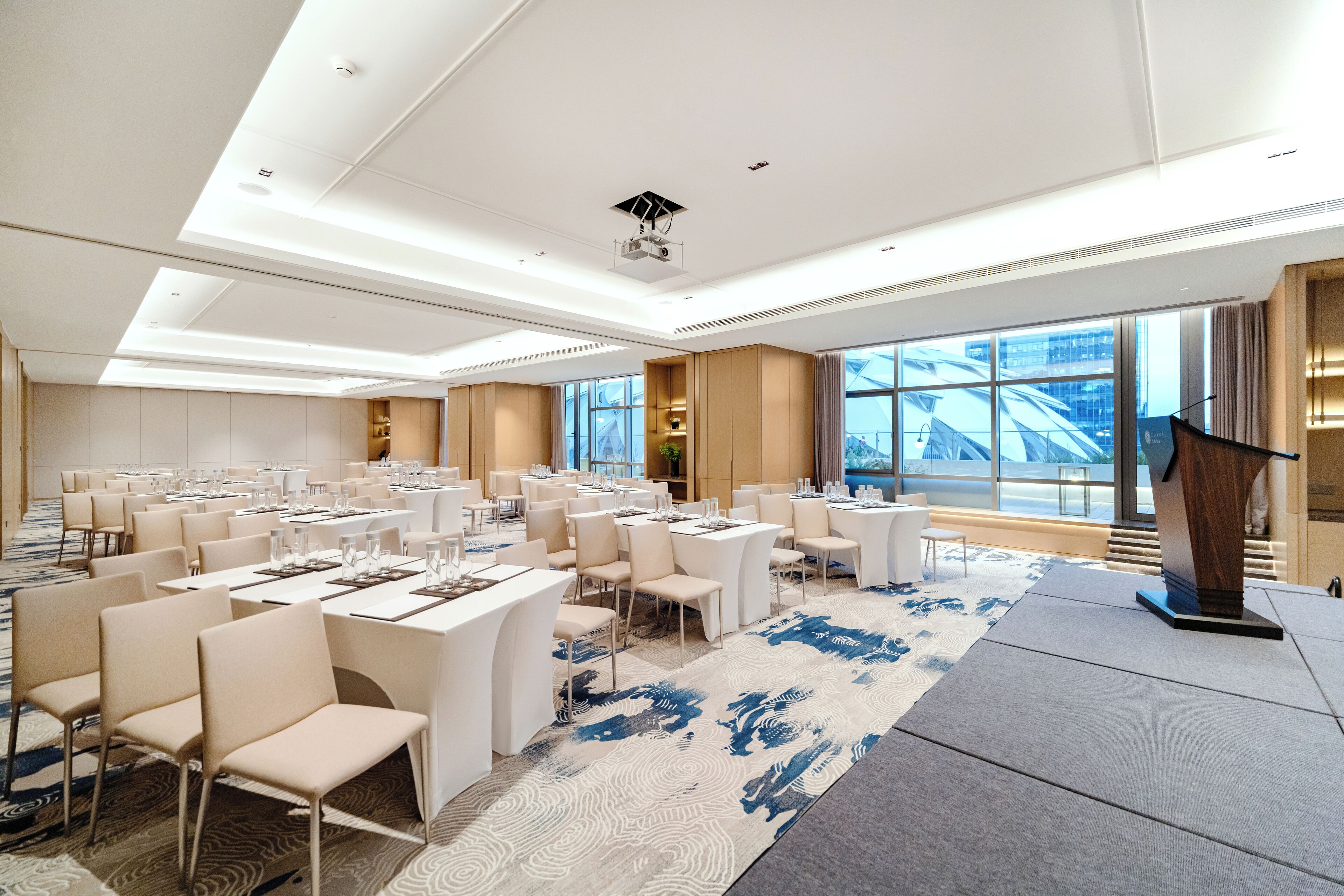 A spacious conference room with multiple tables and chairs, large windows offering a view of the city, and a modern ceiling design.