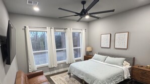 A bedroom with a bed, a ceiling fan, a window with curtains, a bedside table, a lamp, and a brown leather bench.