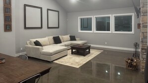 A spacious living room with a sectional sofa, a coffee table, and a dining table. There are three large windows and a fireplace.