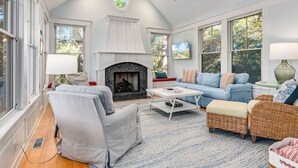 A spacious living room with a fireplace, large windows, and a ceiling fan. - Golf Course view secluded home in the heart of BHI (Bald Head Island)
