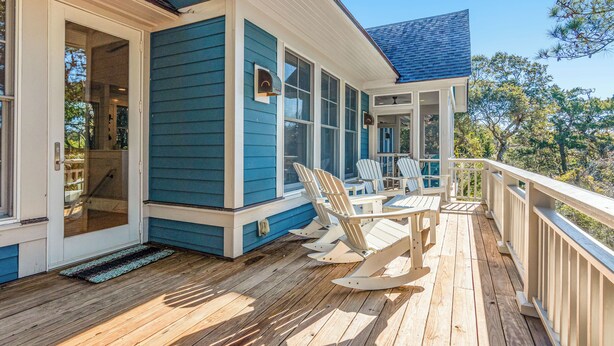 A wooden deck with Adirondack chairs and a table, a covered porch with a railing, and a blue exterior wall. - Golf Course view secluded home in the heart of BHI (Bald Head Island)