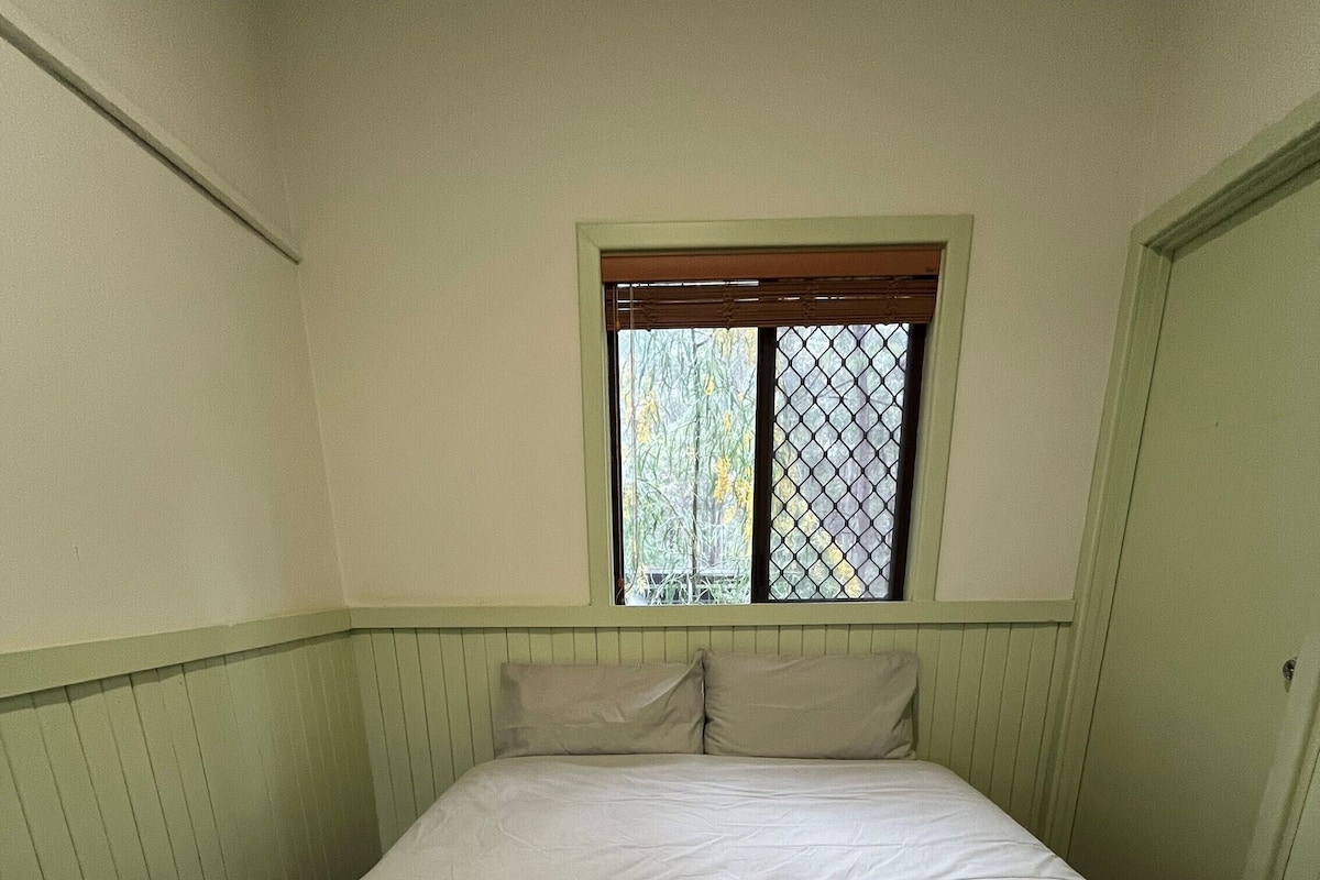 A single bed with a folded white blanket in a room with green walls and a window with a metal screen.