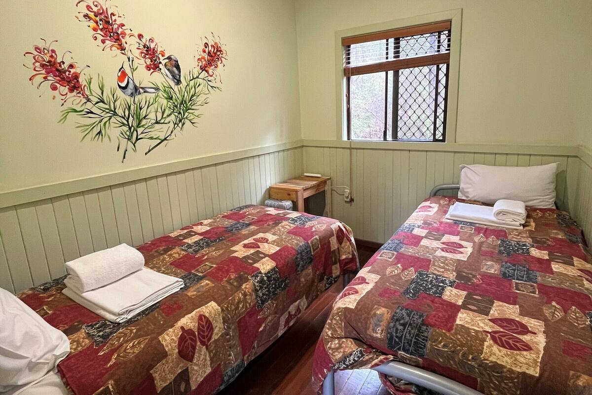 A room with two beds, each with a quilted bedspread and a neatly folded white towel on the bed. There is a window with a wooden frame and a metal screen, and a wall with a floral mural.