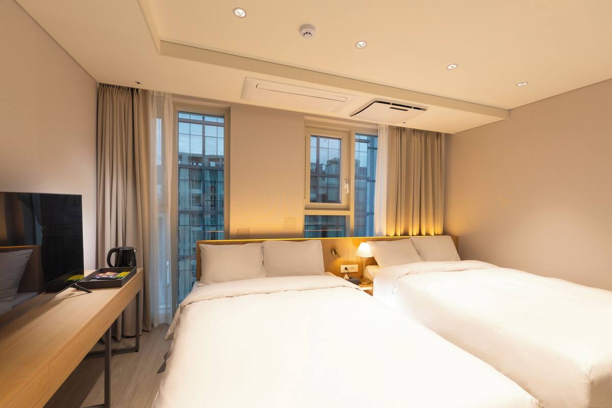 A hotel room with two beds, a desk with a coffee maker, a TV, and large windows with curtains.