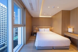 A modern hotel room with a large bed, a nightstand, a desk with a lamp, and a view of a cityscape through a large window.