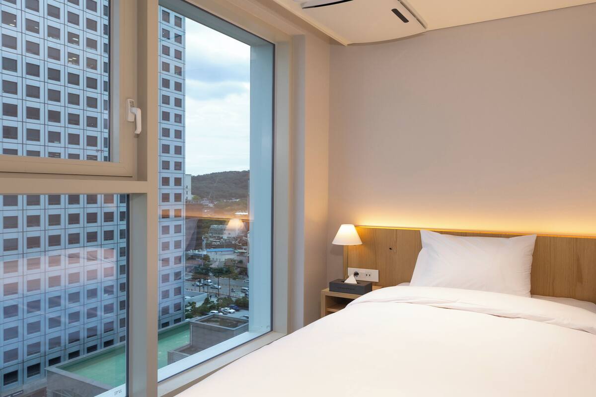 A hotel room with a large window, a bed, a nightstand, and a lamp.