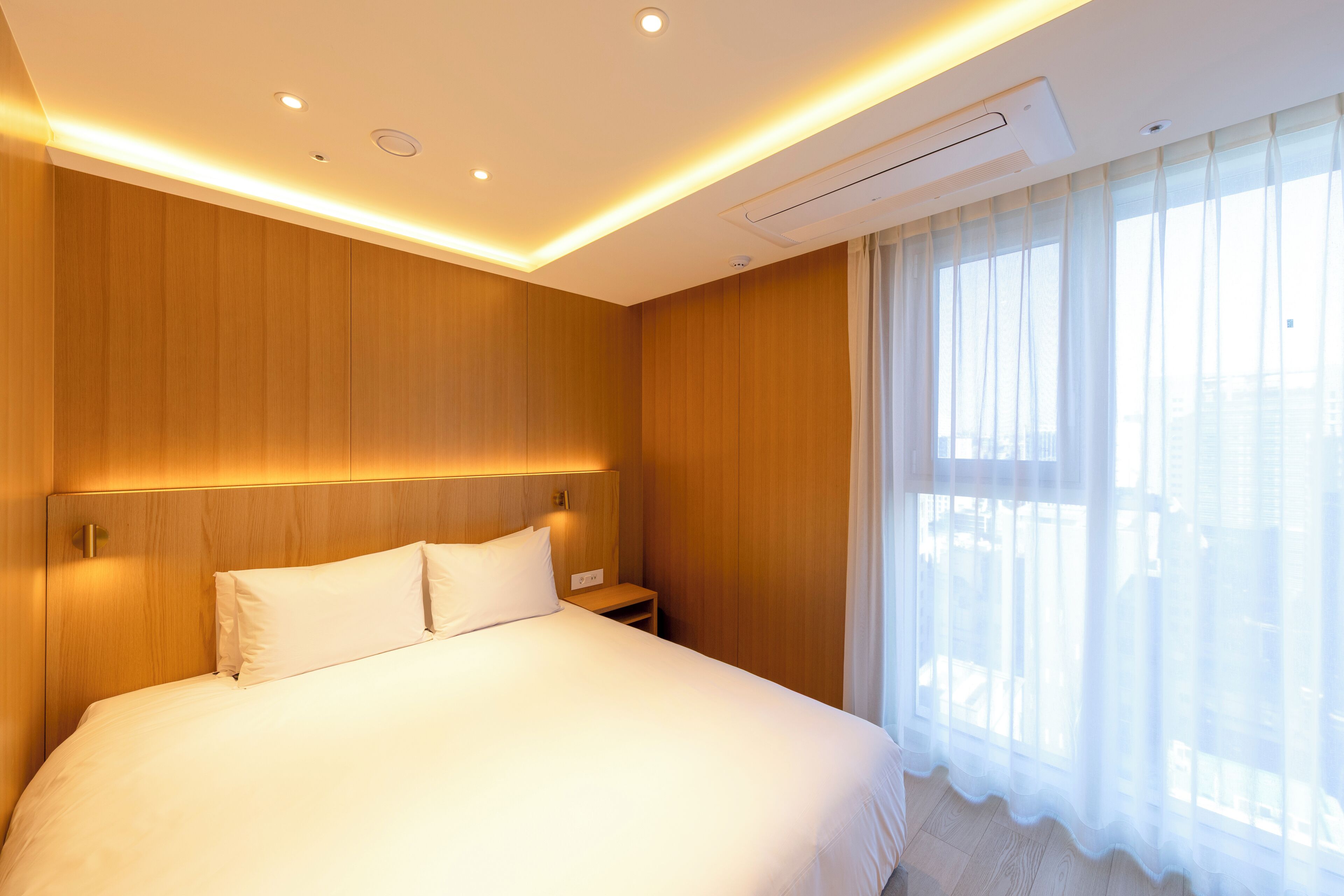 A hotel room with a wooden headboard, a large bed with white linens, a bedside table, and a window with sheer curtains.
