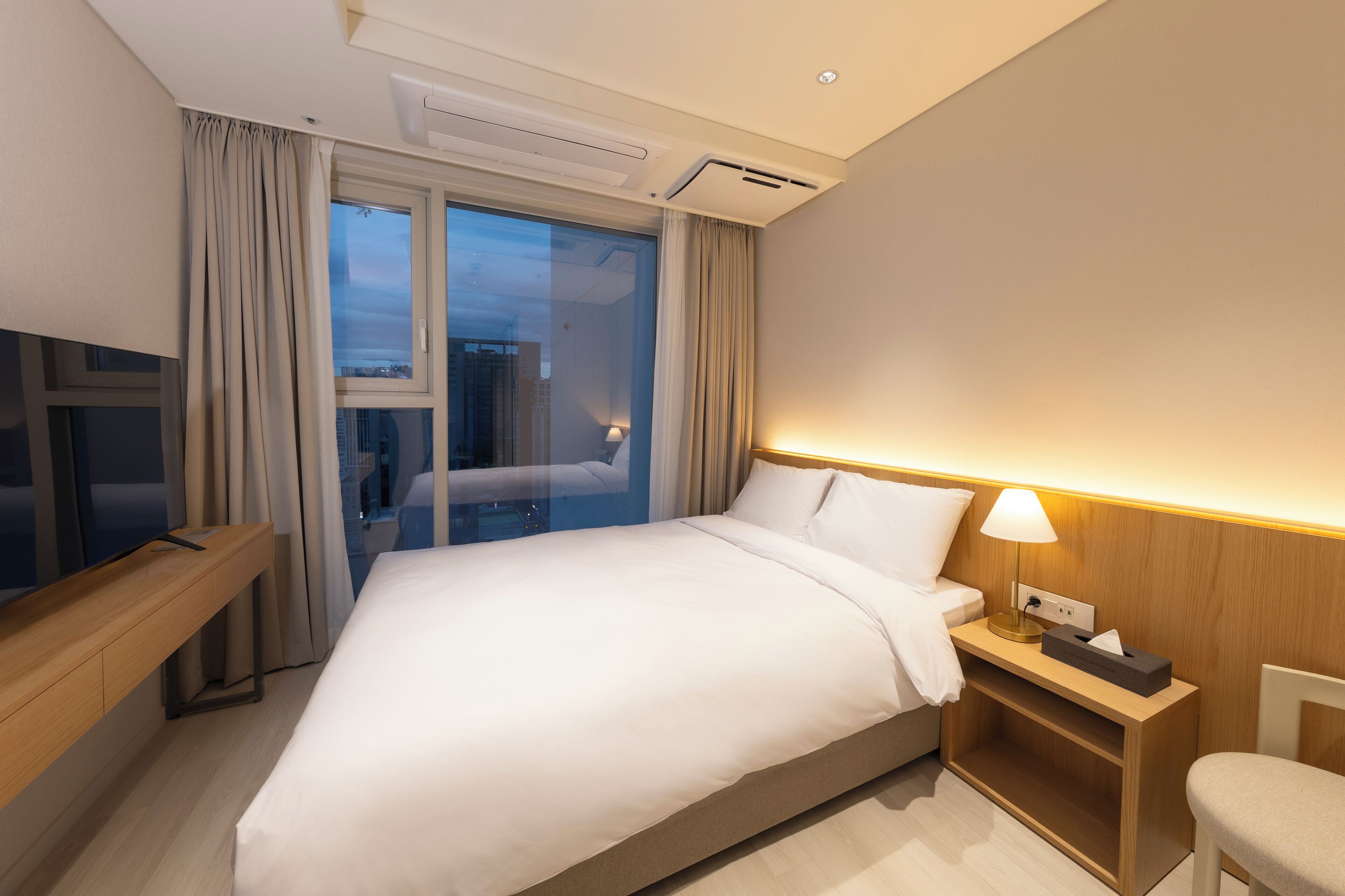 A hotel room with a large bed, a bedside table with a lamp, a TV on a wooden stand, and a window with curtains.