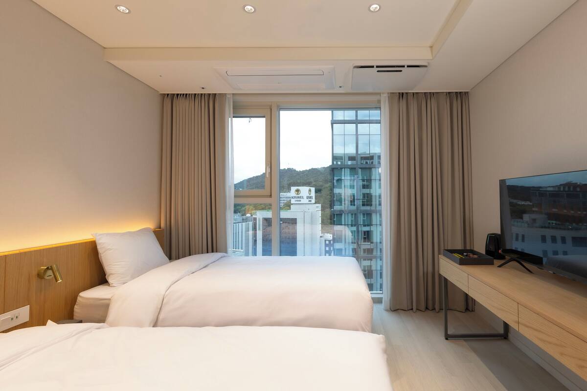 A hotel room with two beds, a TV, and a large window with a city view.