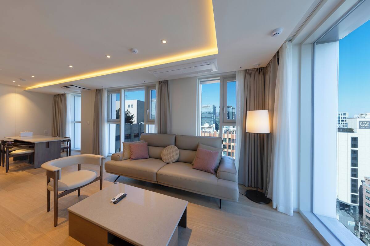 A modern hotel room with a sofa, chairs, a dining table, and a large window offering a city view.