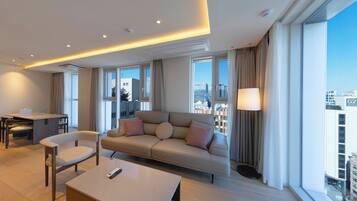 A modern hotel room with a sofa, chairs, a dining table, and a large window offering a city view.