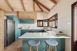A modern kitchen with a large island, wooden beams, teal cabinets, and a stainless steel refrigerator.