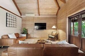 Un salon avec un canapé en cuir marron, une table basse en bois, un téléviseur fixé au mur et une grande fenêtre donnant sur un paysage verdoyant.