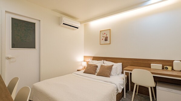 A hotel room with a bed, bedside table, chair, desk, and an air conditioning unit. - NU Hotel (Cheonan)