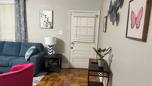 Ein Wohnzimmer mit einem blauen Sofa, einem rosafarbenen Sessel, einem kleinen Tisch mit einer Lampe, einer Topfpflanze und einer Tür mit Wandbildern von Schmetterlingen.