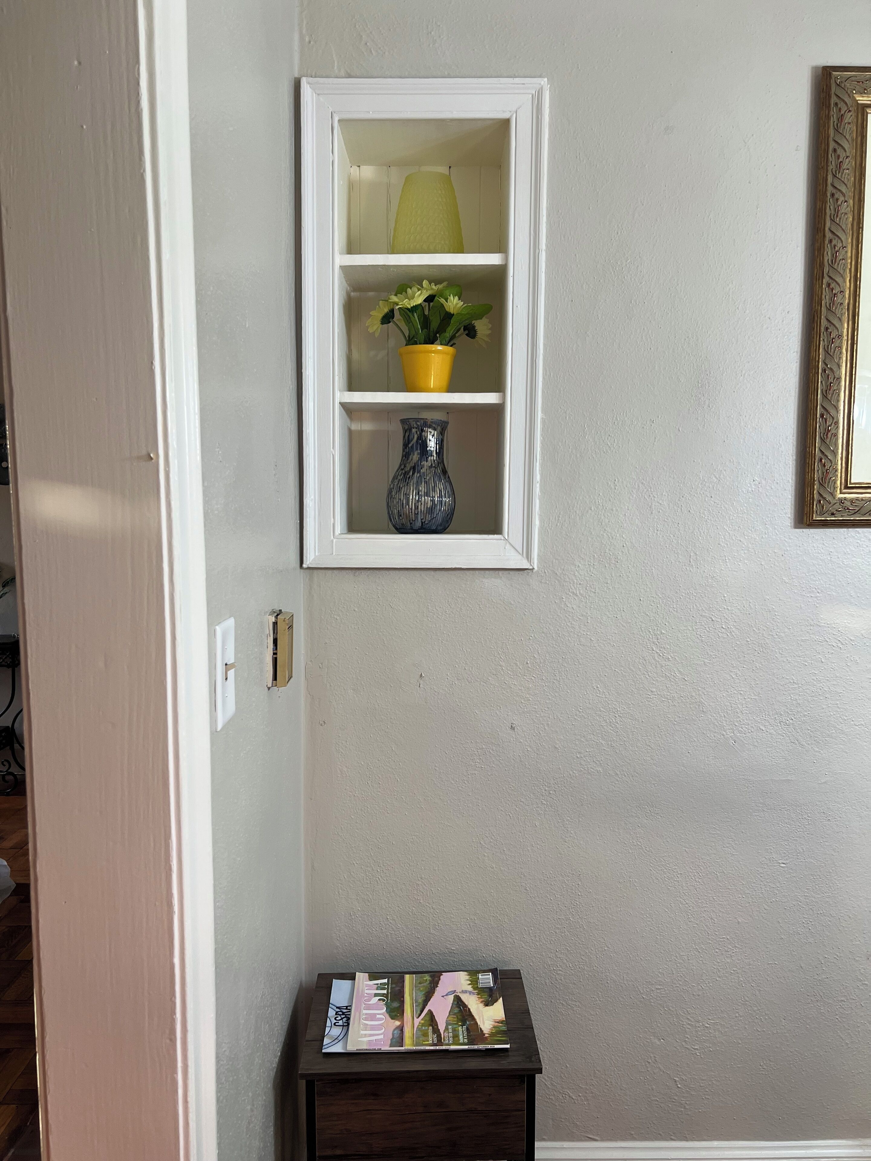 Estante de pared con una maceta amarilla y un jarrón azul, un cuadro enmarcado y una pequeña mesa con revistas.