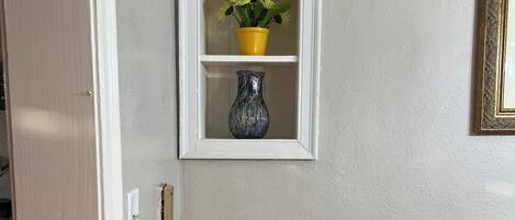 Estante de pared con una maceta amarilla y un jarrón azul, un cuadro enmarcado y una pequeña mesa con revistas.
