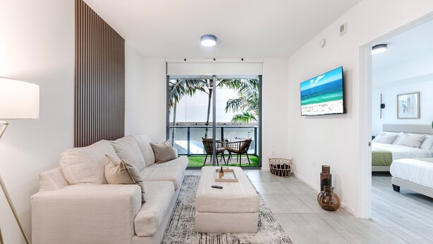 Una sala moderna con sofá, una mesa de centro y televisor. Hay un balcón con vistas a palmeras y al océano.
