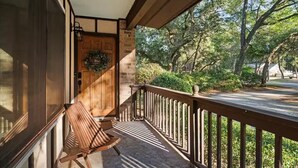 Une véranda en bois avec un banc, une porte ornée d’une guirlande et une vue sur une rue bordée d’arbres.