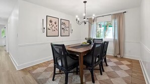 Une salle à manger avec une table en bois, quatre chaises, un lustre et deux œuvres d’art encadrées accrochées au mur.