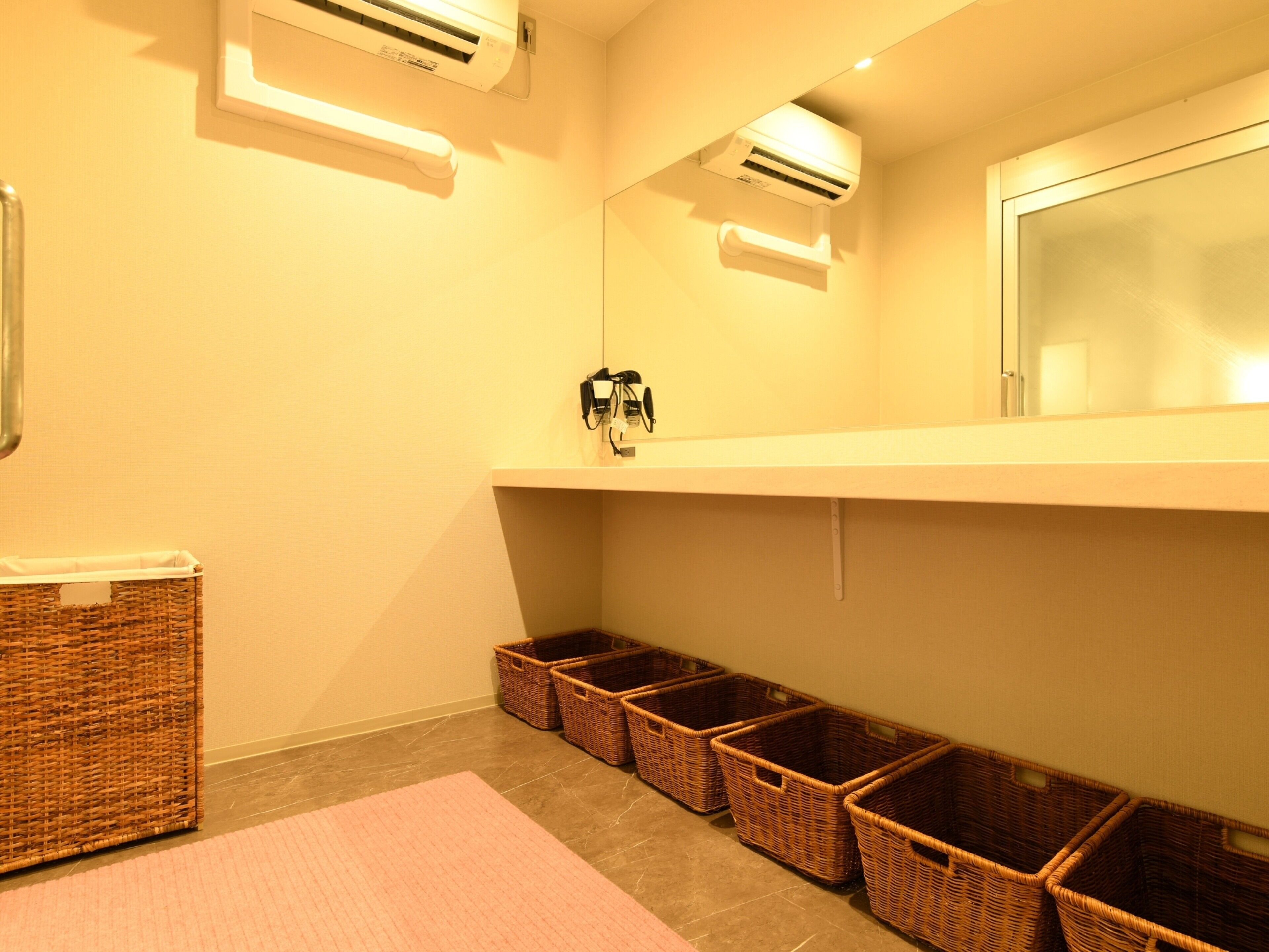 A bathroom with a white sink, mirror, and two wall-mounted air conditioners. There are wicker baskets on the floor and a pink rug.