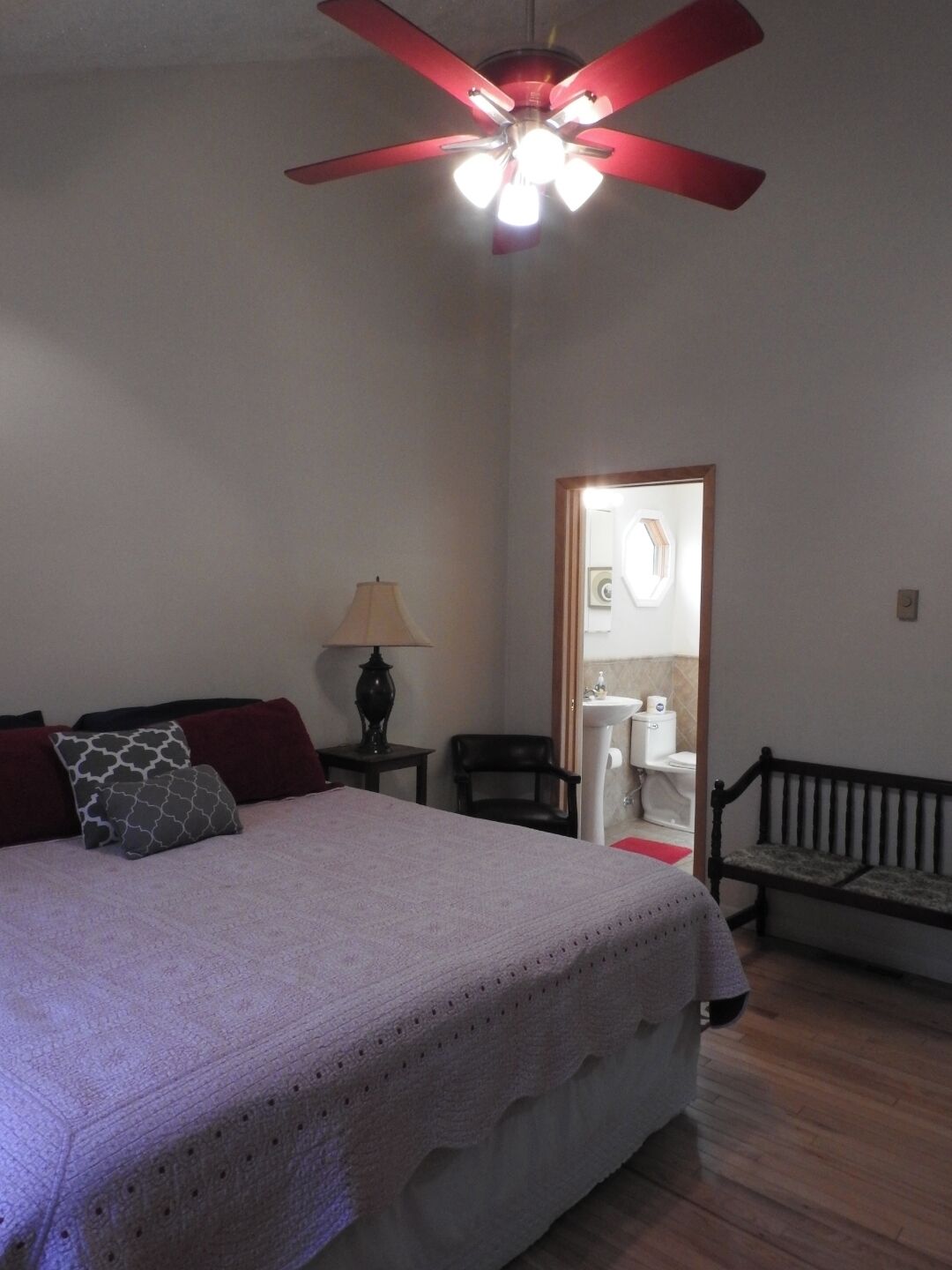 A bedroom with a bed, a ceiling fan, a nightstand, a lamp, a chair, a bench, and a bathroom visible through an open door.