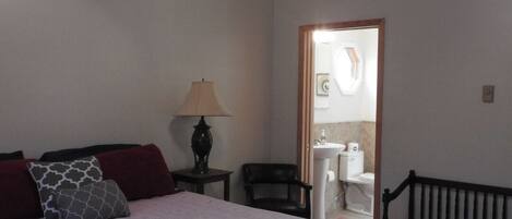 A bedroom with a bed, a ceiling fan, a nightstand, a lamp, a chair, a bench, and a bathroom visible through an open door.