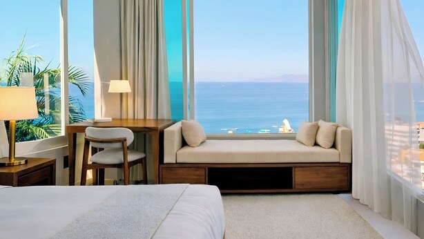 A hotel room with a large window offering a view of the sea, a bed, a desk with a chair, and a small seating area with cushions. - Panoramic Paradise Above Puerto Vallarta. Infinity Pool over Romantic Zone
 (Puerto Vallarta)