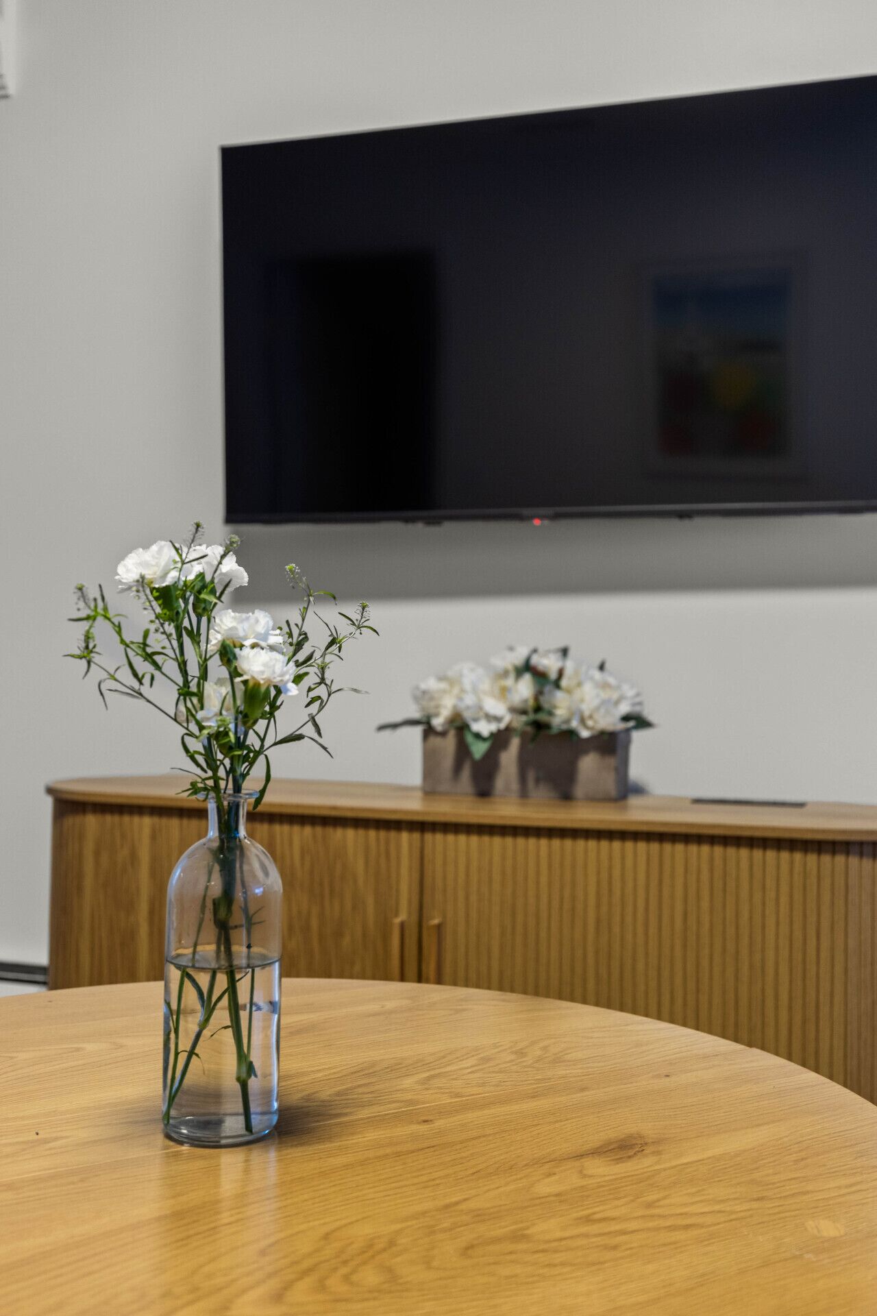 A wooden table with a clear glass vase containing white flowers. In the background, there is a wooden cabinet and a flat-screen TV mounted on the wall.