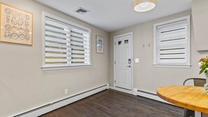 Ein Zimmer mit Holzboden, weißen Wänden und einer Tür mit Schloss. Es gibt ein Fenster mit Jalousien, einen Deckenlüfter und einen kleinen Tisch mit einem Blumenstrauß.