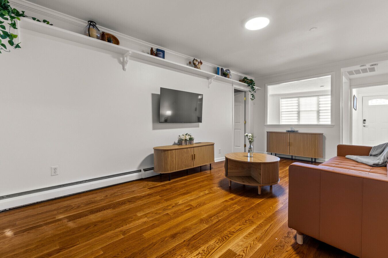 A modern living room with hardwood flooring, a round wooden coffee table, a brown leather sofa, a flat-screen TV mounted on the wall, and a built-in wooden cabinet.