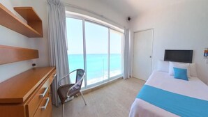 A hotel room with a large window offering a view of the sea, a bed with a blue and white bedspread, a wooden dresser, and a chair. - LUNA SUITE WITH BALCONY AND OCEAN VIEW IN HOTEL ZONE (Cancún)