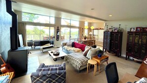 A spacious living room with a large sofa, a coffee table, and a fireplace. There are large windows providing natural light and a view of the outdoors.