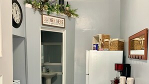 En smal gang med et tekøkken, et ur, et skilt med teksten "GATHER HERE with a grateful heart", samt et køleskab med kurve og stearinlys.